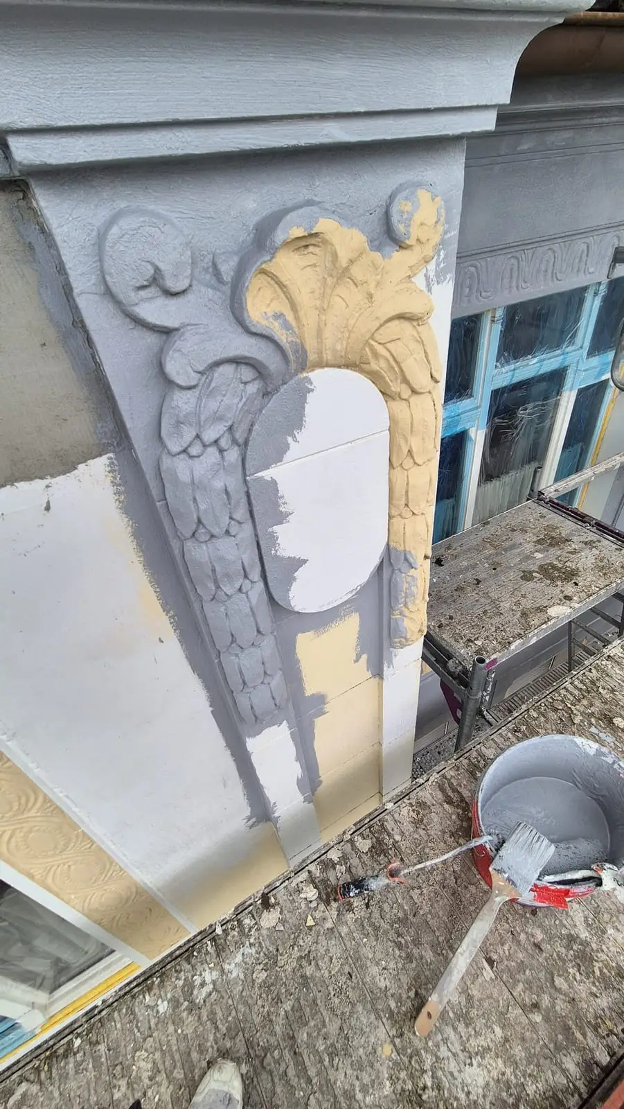 Stuck-Ornament halb im neuen Grau, halb noch im alten Gelb, daneben Pinsel und Eimer